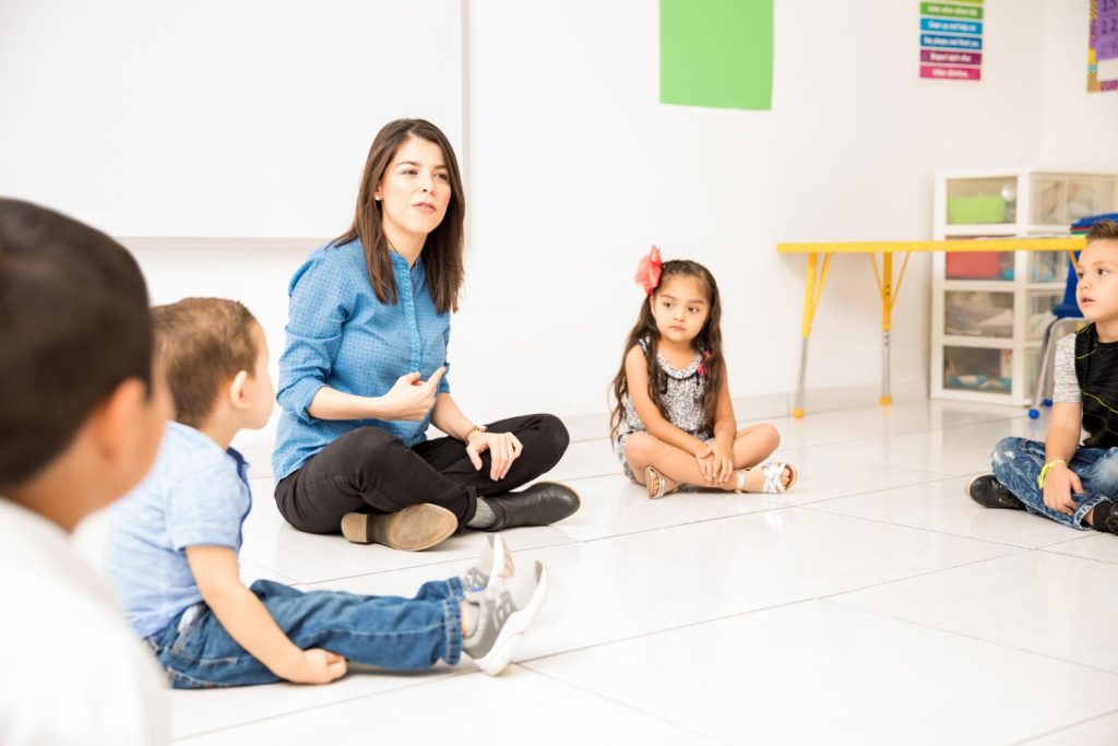 cute-looking-young-woman-teaching-her-preschool-students-while-making-circle-sitting-classroom-floor-1.jpg